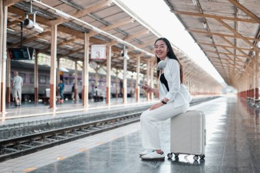 Kulaklıklı ve akıllı telefonlu gülümseyen bir kadın tren istasyonunda bavulun üzerinde oturuyordu, seyahat etmeye hazırdı..