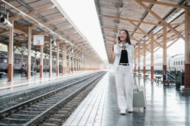 Tren istasyonunda bavullu beyaz elbiseli bir kadın telefonla konuşuyor, seyahat macerasına hazır..