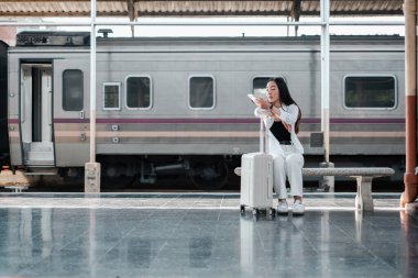 Tren istasyonunda bir bankta bavulla oturan bir kadın, seyahat etmeye hazır. Arka plandaki tren.