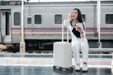 Tren istasyonunda bavuluyla oturan, akıllı telefon kullanan, seyahat etmeye hazır bir kadın. Günlük kıyafetler, modern ulaşım ayarları..