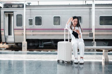 Tren istasyonunda bavullu beyaz elbiseli bir kadın, telefonu kontrol ediyor. Modern seyahat ve iletişim teması.