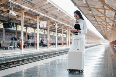 Tren istasyonunda bavullu beyaz elbiseli bir kadın, akıllı telefon kullanıyor. Seyahat ve ulaşım kavramı.