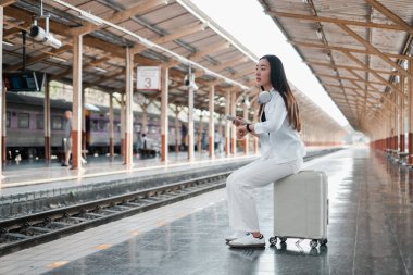 Tren istasyonunda bavulda oturan, kulaklık takan, saate bakan, seyahat etmeye hazır bir kadın..