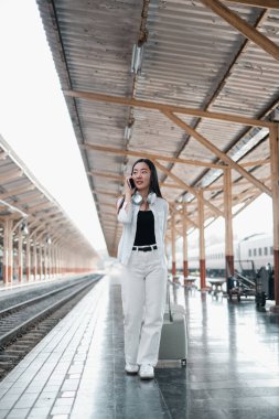 Tren istasyonunda bavullu bir kadın, telefonla konuşuyor, seyahat macerasına hazır. Gündelik tarz, modern yolculuk..