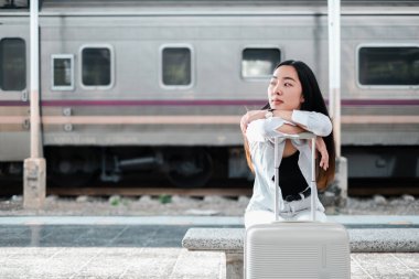 Genç bir kadın tren istasyonunda bankta oturuyor, bavulları yanında, seyahat, beklenti ve şehir keşiflerini somutlaştırıyor..
