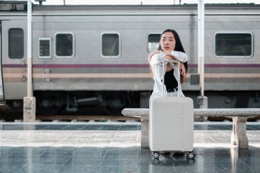 Genç bir kadın tren istasyonunda bavuluna yaslanır ve yolculuğa hazırdır. Tren arka planda görünür..