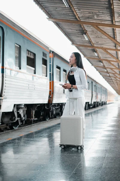 Beyaz elbiseli şık bir kadın tren istasyonunda bavuluyla yolculuk için hazır bekliyor. Modern ulaşım ve seyahat kavramı.