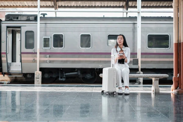 Tren istasyonunda oturan bavullu bir kadın, telefonunu kullanıyor. Seyahat, bekleme ve modern ulaşımı temsil eder..