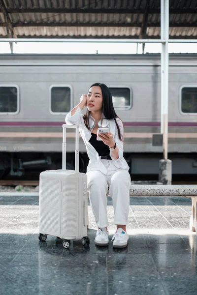 Tren istasyonunda elinde bavulla oturan, akıllı telefon kullanan, sıradan giyinmiş, ulaşımı bekleyen bir kadın..