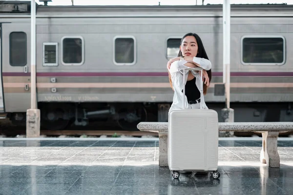 Genç bir kadın tren istasyonunda bavuluna yaslanır ve yolculuğa hazırdır. Tren arka planda görünür..