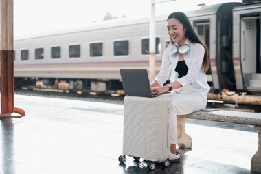 Tren istasyonunda bavulların üzerinde otururken dizüstü bilgisayar kullanan gülümseyen kadın, seyahat ve uzak iş yaşam tarzını benimsiyor..