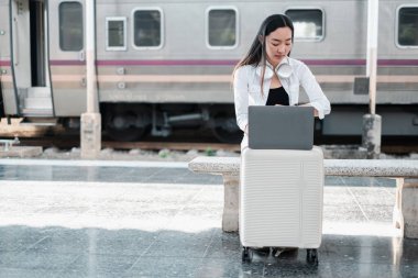 Genç bir kadın, bir tren istasyonunda bavulunun üzerine oturmuş dizüstü bilgisayarıyla çalışıyor. Seyahat etmeyi ve uzaktan çalışmayı simgeliyor..