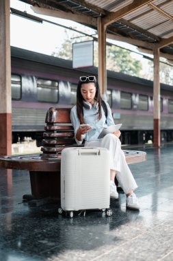 Genç bir kadın gezgin, bir tren istasyonunda bavuluyla bankta oturuyor, akıllı telefonu ve tabletini kullanıyor..