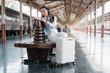 Genç bir kadın tren istasyonunda oturmuş kitap okuyor, bavulları yanında, seyahat ve keşfi modern bir ortamda somutlaştırıyor..