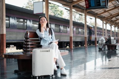 Tren istasyonunda bavuluyla oturan, gülümseyen ve rahatlamış, seyahat macerasına hazır bir kadın..