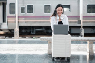 Kulaklıklı gülümseyen kadın tren istasyonunda dizüstü bilgisayar kullanarak seyahat ve uzaktan çalışmayı kucaklıyor..