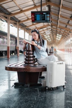 Tren istasyonunda bavuluyla oturan, kitap okuyan, seyahat ve keşfi simgeleyen bir kadın..