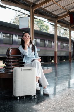 Tren istasyonunda bavuluyla oturan bir kadın, seyahatten önce biraz dinlenmenin tadını çıkarıyor..