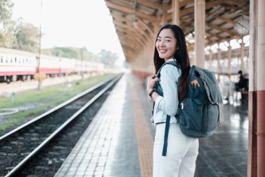 Sırt çantalı gülümseyen kadın tren istasyonunda bekliyor, yolculuğa hazır. Parlak, güneşli bir gün. Trenler arka planda..