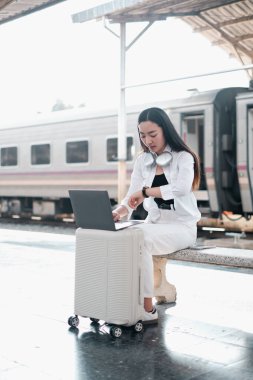 Genç bir kadın tren istasyonunda bavulunun üzerine oturmuş, trenini beklerken dizüstü bilgisayarıyla çalışıyor..