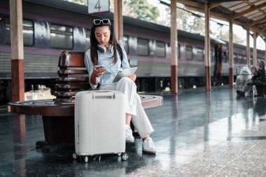 Tren istasyonunda elinde bavulla oturan, akıllı telefon ve tablet kullanan, seyahate hazırlanan bir kadın..