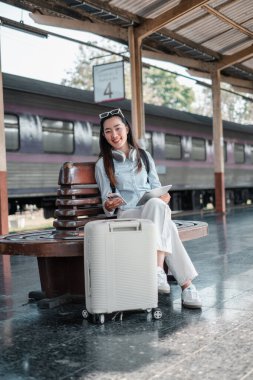 Tren istasyonunda bavul ve tabletle oturmuş gülümseyen bir kadın, seyahat macerasına hazır..