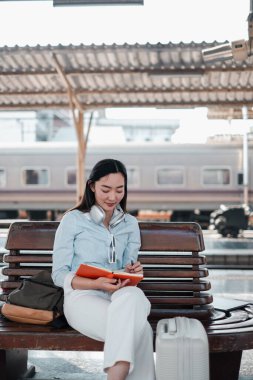 Tren istasyonunda bankta oturan bir kadın, boynunda kulaklıklar ve yanında bavullarla bir deftere yazıyor..