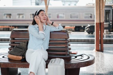 Kulaklıkla müzik dinleyen genç bir kadın, tren istasyonunda bir bankta oturuyor, bavul ve sırt çantasıyla..