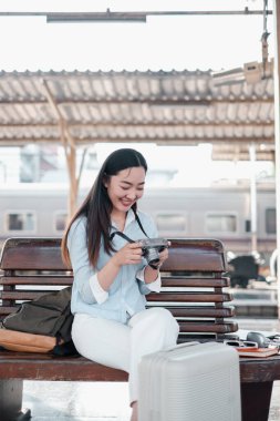 Genç bir kadın tren istasyonunda bir bankta oturuyor, elinde bir kamera ve gülümsüyor, yanında bavullarıyla, seyahat etmeye hazır..