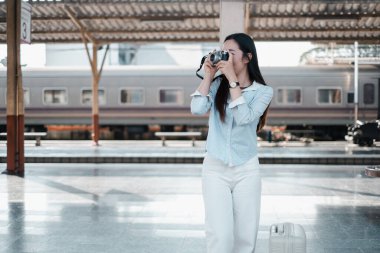 Klasik kameralı genç bir kadın tren istasyonunda duruyor, anları kaydetmeye hazır. Yanında bir bavul var..