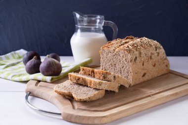 Beyaz bir masada, üzerinde taze dilimlenmiş ekmek olan bir tahta, bir sürahi süt ve olgun incir, mavi arka planda.