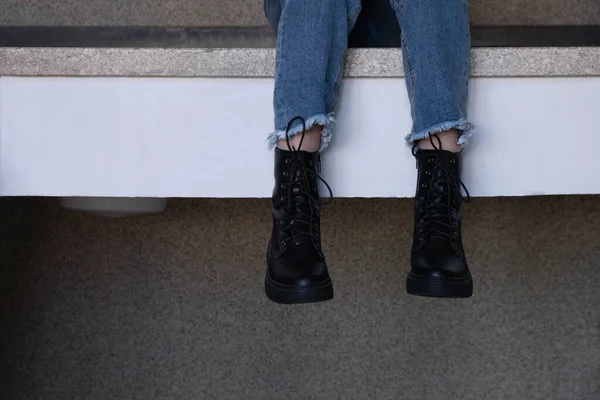 Bacaklar, siyah botlar içinde, korkuluklardan aşağı itilmiş, sallanıyor.