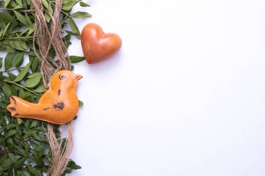 Metalden yapılmış yaşlı bir kuş, taştan bir kalbin yanında ve yeşil yaprakların üzerinde, beyaz arka planda, üst görünümde.