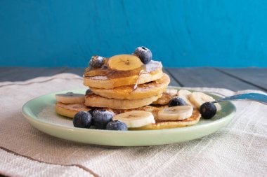 Bal serpiştirilmiş gözlemeler, böğürtlenler ve doğranmış muzlar bir tabakta, masaya pudra şekeri serpiştirilmiş.