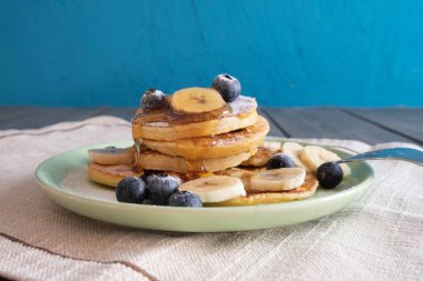 Taze gözlemeler, bal, böğürtlenler ve dilimlenmiş muzlarla dolu bir tabakta, üzerine pudra şekeri serpiştirilmiş, masanın üzerine