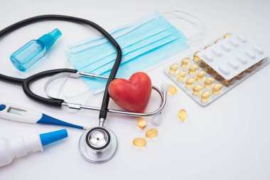 Maske, vitaminler, stetoskop, kırmızı kalp, antiseptik, termometre, masanın üstünde beyaz bir arka plan.