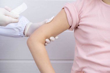 Doktor çocuğun eline bir enjeksiyon yaptı, enjeksiyon yerini pamuklu bezle sıktı ve beyaz bir arka plana alçı uygulamak istiyor.