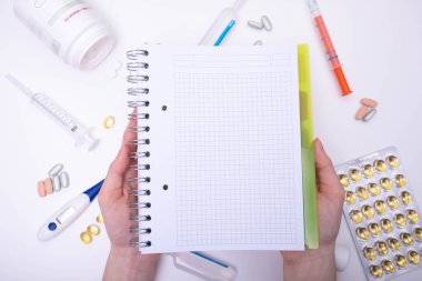 Defter ellerinde, termometrenin, şırınganın, hapların ve çeşitli tıbbi malzemelerin arkasında beyaz bir arka planda duruyor.