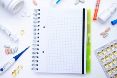 Defter masanın üzerinde duruyor. Yanında da bir termometre, şırınga, haplar ve çeşitli tıbbi malzemeler var. Beyaz bir arka planda.