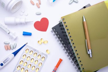 Not defterleri masanın üzerinde, ve beyaz arka planda termometrenin, şırınganın, hapların ve çeşitli tıbbi malzemelerin yanında.