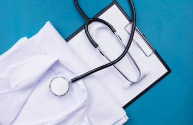 Steteskop, beyaz doktor üniforması katlanmış ve mavi arka planda, masanın üzerinde yatıyor.