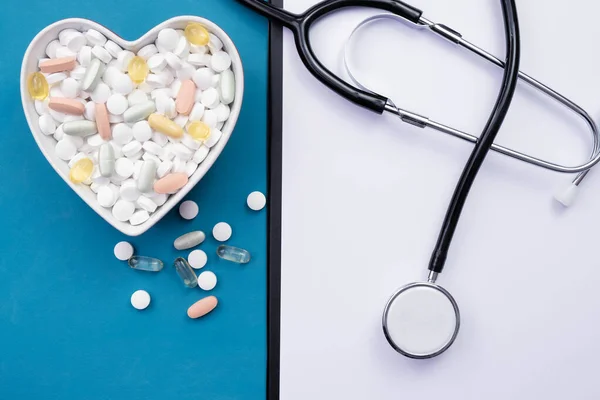 heart shaped cup full of pills on the table, stethoscope and notepad for behind, on a blue background