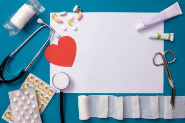 Bandajlar, makas, kırmızı kalp, steteskop, haplar ve beyaz bir kağıt parçası masanın üzerinde, mavi arka planda.