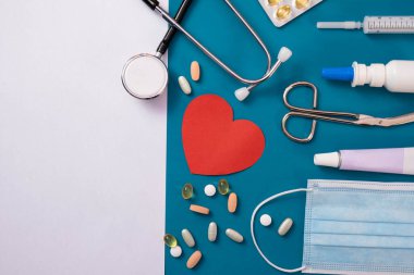 kırmızı kalp, maske, bandaj, makas, merhem, steteskop, haplar ve beyaz bir kağıt parçası masanın üzerinde, mavi bir arka planda.