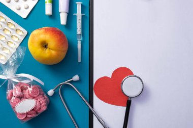 Şırınga, kesilmiş kalp, stetoskop, not defteri, kalp, elma, vitamin, hap, maske mavi arka planda, masanın üzerinde.
