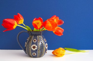 Mavi bir sürahide kırmızı laleler açıyor. Beyaz bir masanın üzerinde bir desen duruyor. Ve bir lale yakınlarda, mavi arka planda yatıyor.