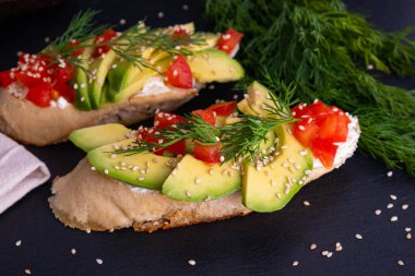 İştah açıcı kanepeler, taze avokado dilimleri ve olgun domates dilimleri bir parça baget üzerine serpilmiş ve susam tohumları serpiştirilmiş, siyah arka plan