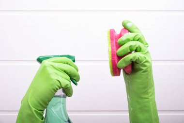Yeşil eldivenli eller, beyaz arka planda banyoyu temizlemek için sünger ve deterjan tutuyor.