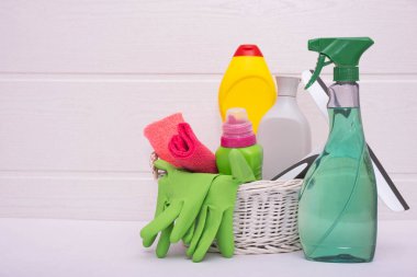 Beyaz arka planda temizlik sıvıları, süngerler, fırçalar, paçavralar ve eldivenlerle dolu bir sepet.