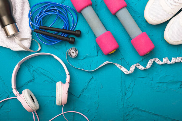 sport at home - white sneakers, dumbbells and jump rope, measurement tape, towel and water bottle, headphones lie on a blue textured background